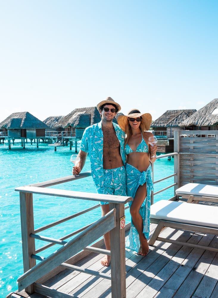 a man and woman standing on a dock in front of water with thatched huts