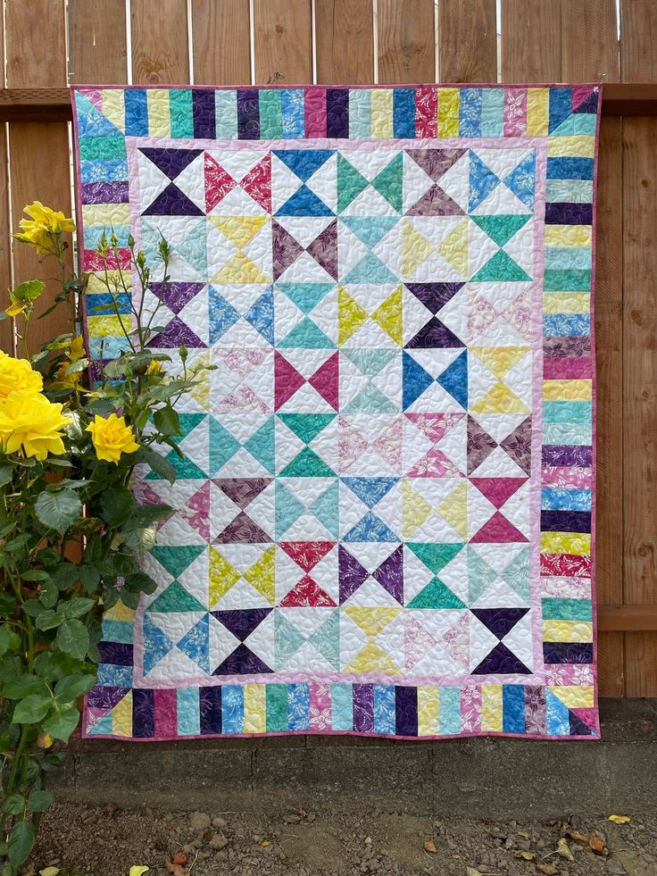 a colorful quilt next to yellow flowers in front of a wooden fence