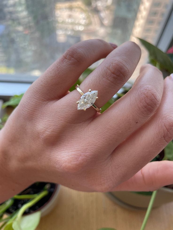 a woman's hand with a diamond ring on top of her finger, next to a potted plant