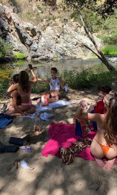 several people sitting on the ground near a river with towels and drinks in their hands