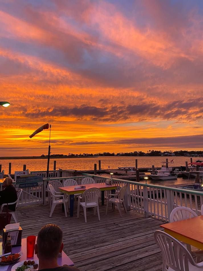 people are sitting at tables on the dock watching the sun go down over the water