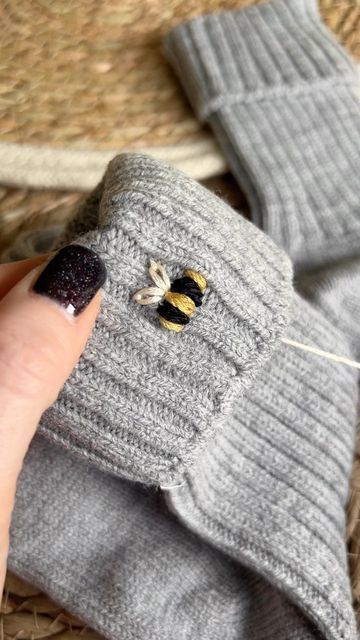 a hand with a black nail polish holding onto a gray knitted mitt that has a bee on it