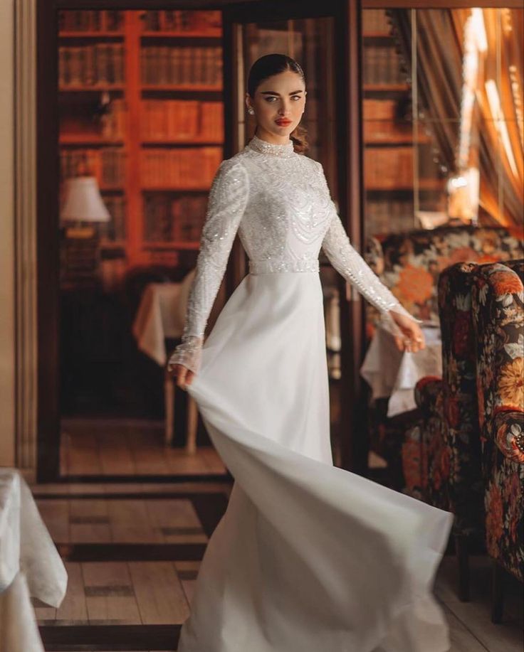 a woman in a white dress is standing near a chair and bookshelf, wearing a long sleeved wedding gown