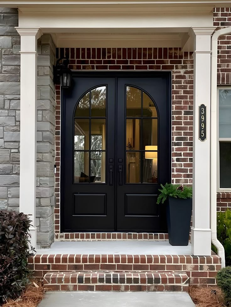 a black front door with two potted plants on the steps and brick building behind it