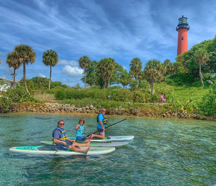 three people on paddle boards in front of a light house with palm trees and blue sky