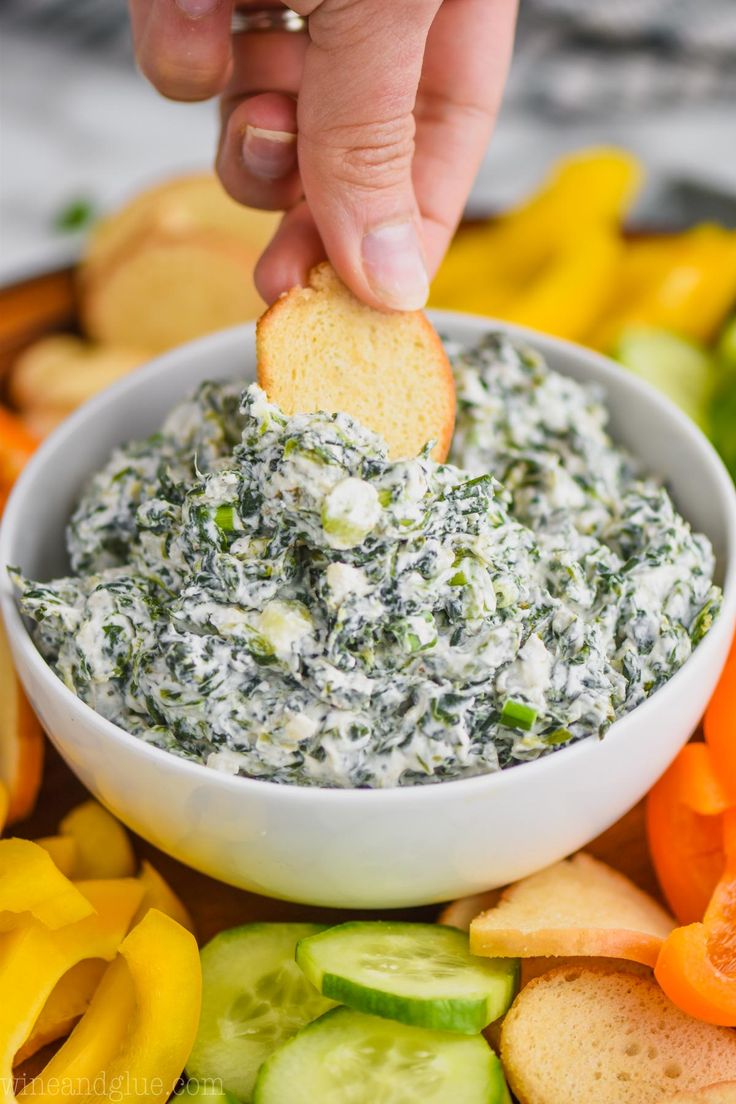 a hand dipping a cracker into a bowl of spinach dip surrounded by vegetables