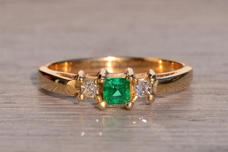 The Tule River: Natural Emerald and Diamond Three Stone Ring in Yellow Gold. At the heart of this elegant ring lies a striking 0.20 carat square emerald cut natural emerald, radiating a medium, very slightly bluish-green hue with strong saturation. Flanking the center gem are square modified brilliant cut (princess cut) natural diamonds, boasting an approximate total weight of 0.14 carats by measurement. Meticulously crafted in 14 karat yellow gold, the ring, sized at 7, exudes timeless charm an Heart Cut Green Emerald Ring For Formal Occasions, Green Heart Cut Emerald Ring For Formal Occasions, Formal Green Heart Cut Emerald Ring, Elegant Heart Cut Emerald Ring Gift, Elegant Heart Cut Green Emerald Ring, Elegant Rectangular Emerald Ring With Prong Setting, Elegant Rectangular Emerald Ring With Center Stone, Elegant Green Emerald Heart Cut Ring, Elegant Green Heart Cut Emerald Ring