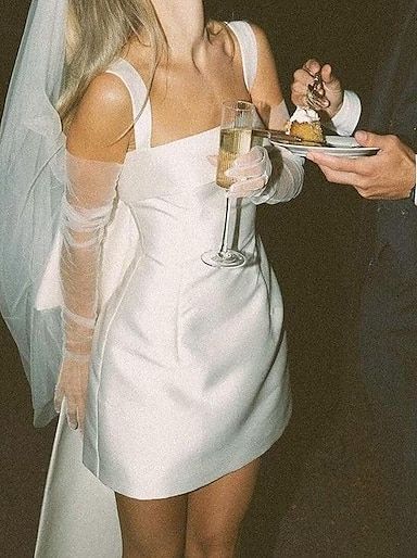 a woman in a white dress holding a plate with food on it while standing next to a man wearing a suit and tie