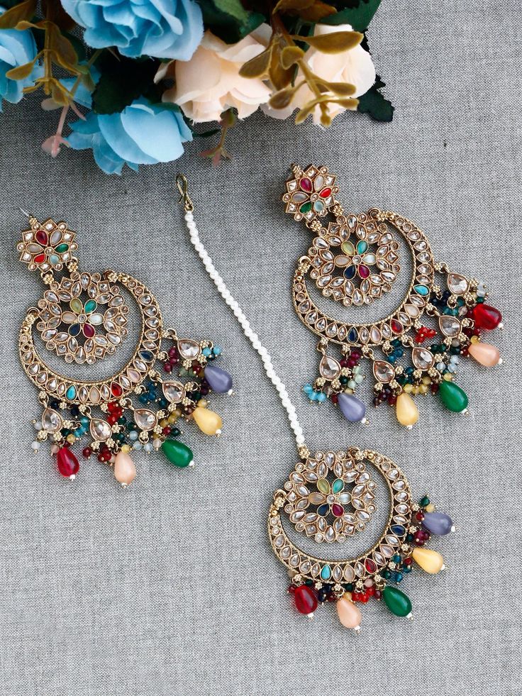 "Add a vibrant burst of colors to your ensemble with our stunning Multicoloured Navratan Maang Tikka and Earring Set. Inspired by the essence of Navratan, which means \"nine gems,\" this ensemble features a harmonious combination of colorful gemstones that exude a joyful and celebratory spirit. The centerpiece of this set is the maang tikka, a traditional forehead adornment that adds a regal touch to your look. It showcases an array of gemstones, including ruby red, emerald green, sapphire blue, topaz yellow, and more, each representing one of the nine gems. The intricate design and delicate craftsmanship reflect the cultural heritage and traditional significance of Navratan jewelry. Coordinating with the maang tikka, the earrings complete the ensemble with their vibrant beauty. They featu Traditional Multicolor Chandbalis With Latkans, Festive Multicolor Chandbali Jhumkas, Multicolor Chandbali Jhumkas For Festive Occasions, Multicolor Traditional Jhumkas For Celebrations, Traditional Multicolor Jhumkas For Celebrations, Multicolor Peacock Design Jhumkas For Festivals, Multicolor Tilla Jhumkas For Navratri, Multicolor Jhumkas For Navratri Festival, Multicolor Jhumkas For Eid Festival