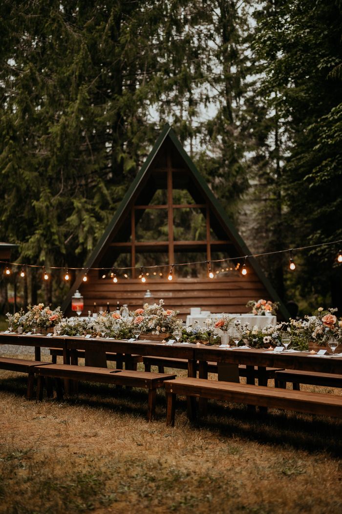 a long table with flowers and candles on it in front of a wooden cabin surrounded by trees