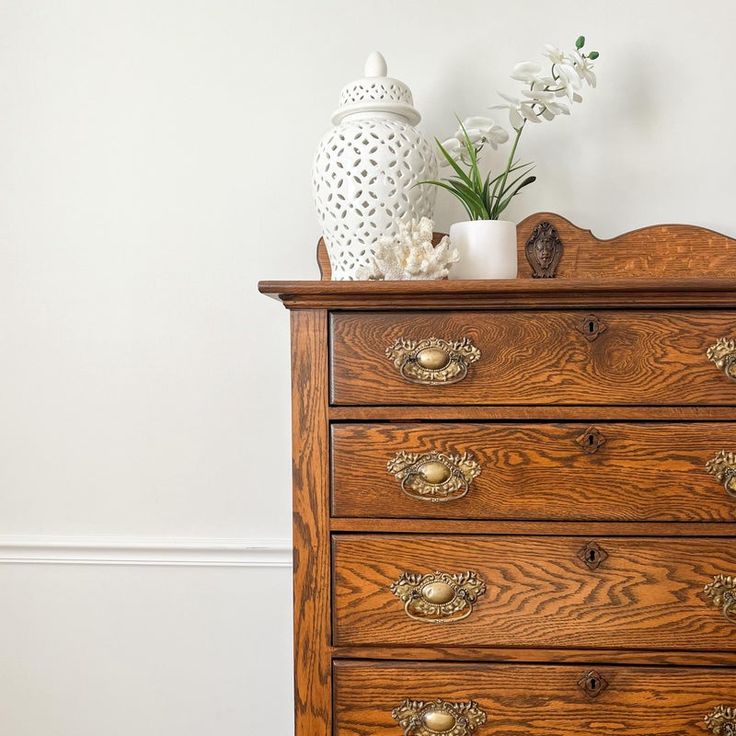 a wooden dresser with flowers and a vase sitting on it's top, against a white wall