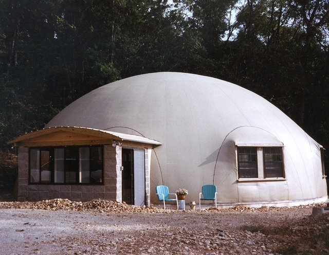 a round building with two blue chairs in front of it and trees behind the building