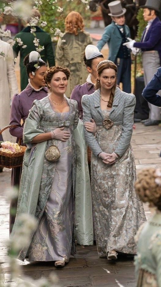 two women dressed in period costumes walking down the street with other people behind them and one woman holding an umbrella