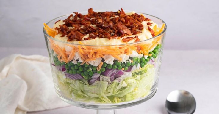 a salad in a glass dish with bacon and peas on top next to a spoon