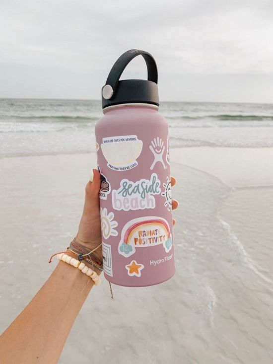 a hand holding a pink water bottle with stickers on it at the ocean shore