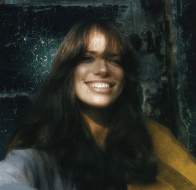 a woman with long hair smiling and wearing a yellow scarf over her shoulders, in front of a grungy wall