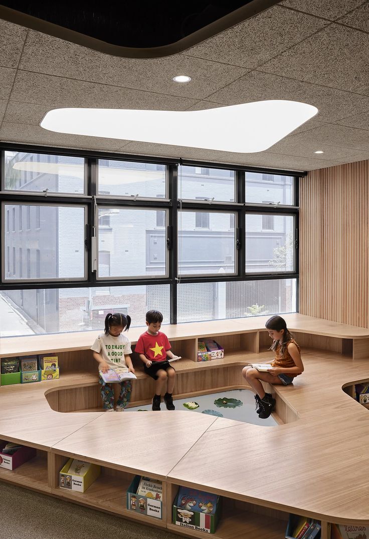 three children sitting on benches in a library with bookshelves and windows overlooking the city