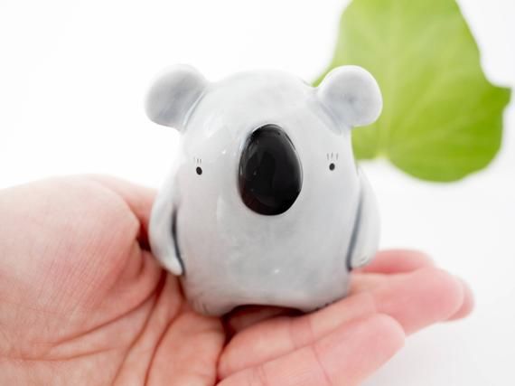 a small ceramic koala bear sitting on top of a persons hand next to a green leaf