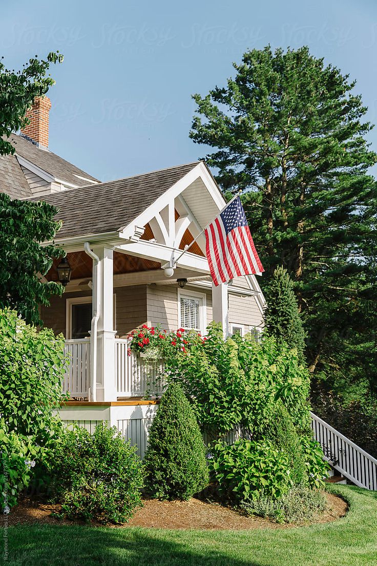 a house with an american flag on the porch by some bushes and trees in front of it