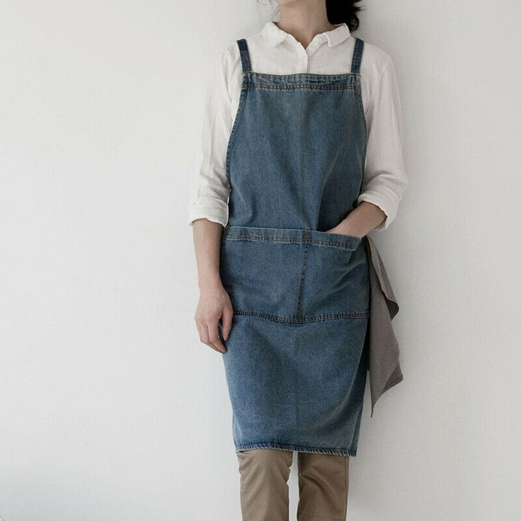 a woman standing in front of a white wall wearing an overall dress and brown boots