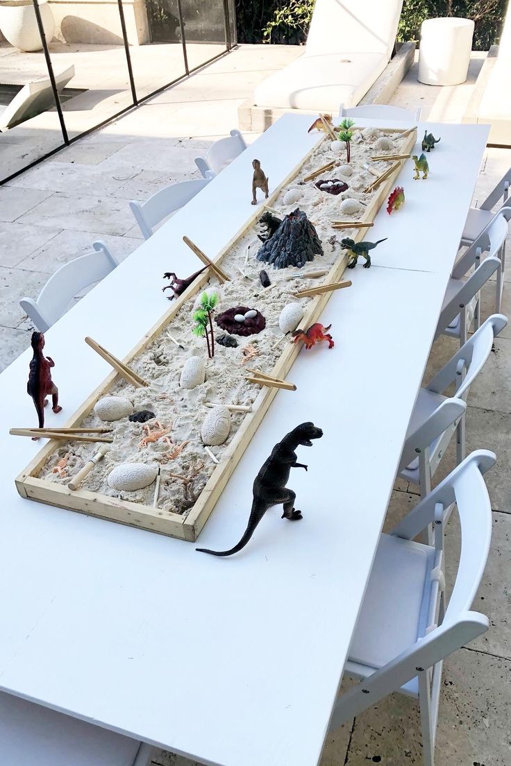 a long table with toy dinosaurs and sand in the shape of a boat on it