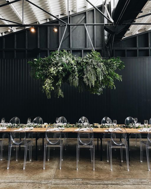 a long wooden table with clear chairs and greenery hanging from it's ceiling