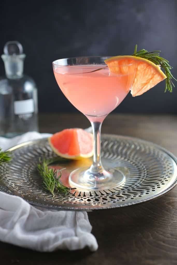 a pink drink with orange slices and rosemary garnish on a silver plate next to a bottle of gin