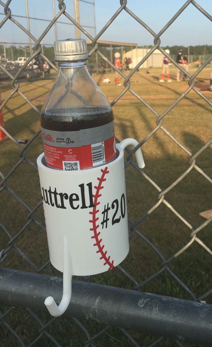 a plastic bottle with a baseball on it hanging from a chain link fence in front of a baseball field