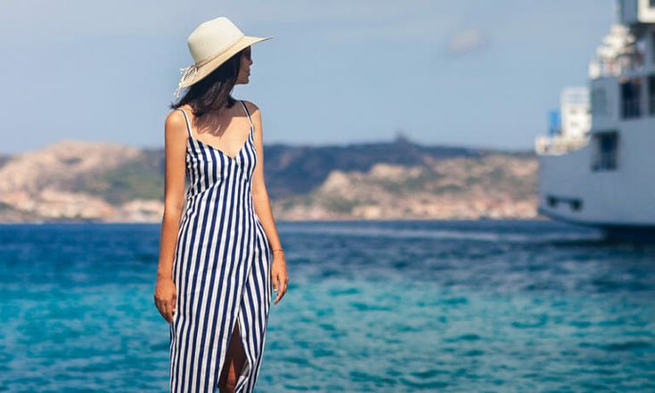 a woman in a striped dress and hat standing on the beach looking out at the water