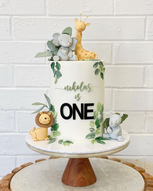 a white cake decorated with jungle animals and greenery on a wooden stand in front of a brick wall