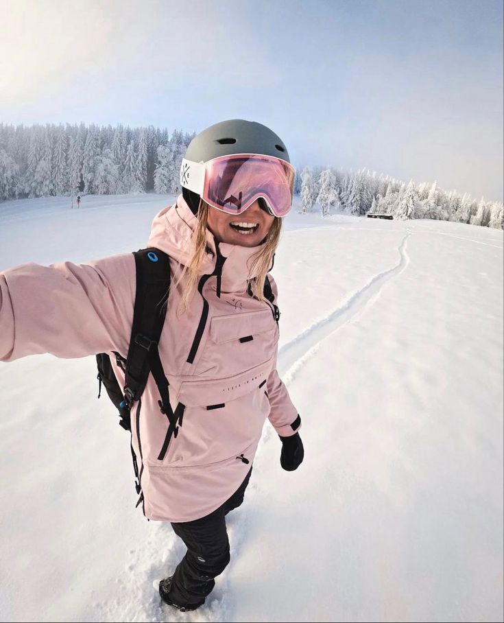 a woman in pink jacket and goggles on snow covered ground with trees behind her