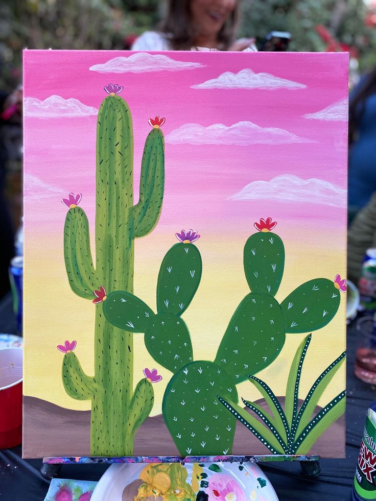 a painting of a cactus with pink sky and clouds in the background is displayed on a table