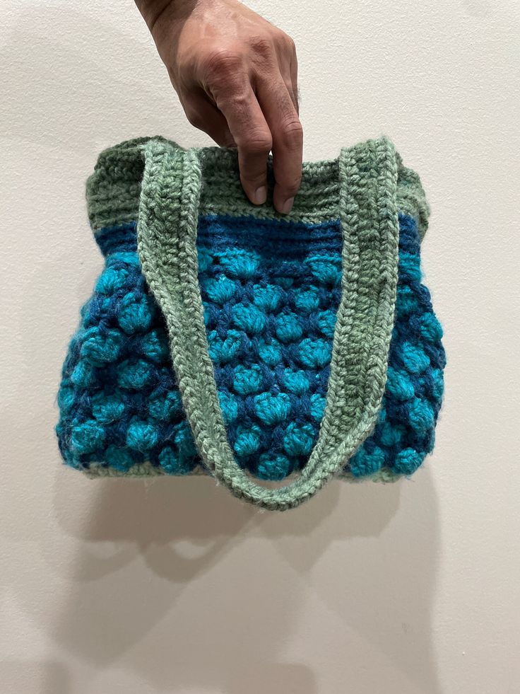 a hand is holding a crocheted blue and green purse on a white wall