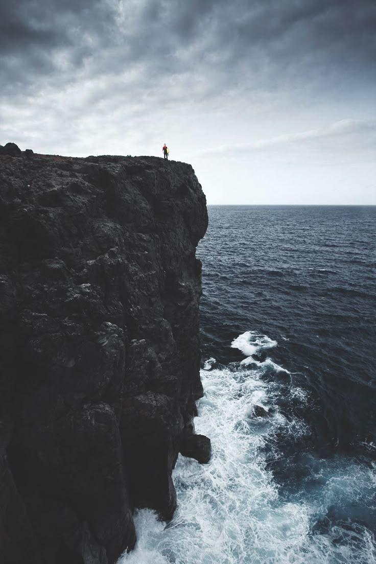 a person standing on top of a cliff next to the ocean under a cloudy sky