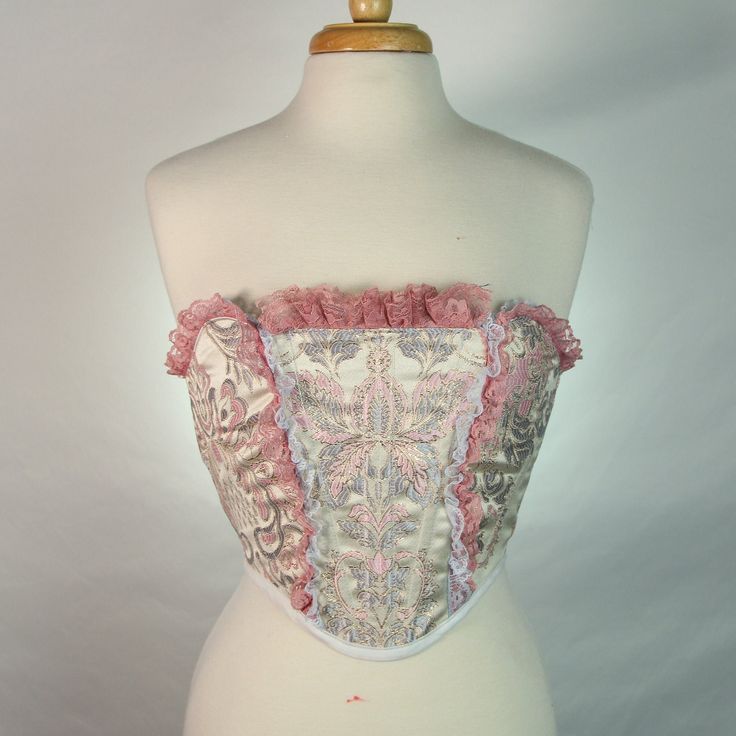 Over-bust strapless corset with flexible boning and mauve lacing in back. Ivory, pastel and gold victorian style silk with mauve and powder blue accent ruffle lace.  No sizing options available because this corset is made to order based on your proportions.  Mannequin's measurements - Waist: 27" Bust: 37" Feminine Strapless Corset With Lace Trim, Feminine Strapless Lace Corset, Strapless Feminine Corset With Lace Trim, Strapless Lace Corset In Feminine Style, Elegant Overbust Bodice With Ruffles, Pink Lace Trim Underbust Corset, Pink Lace Underbust Corset, Pink Underbust Corset With Lace Trim, Pink Lace Corset With Boned Bodice