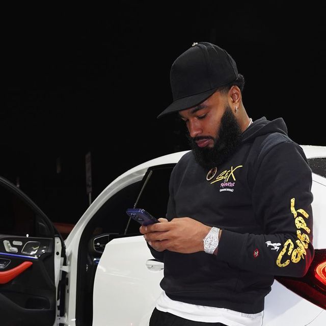 a man standing next to a white car looking at his cell phone while wearing a black hat