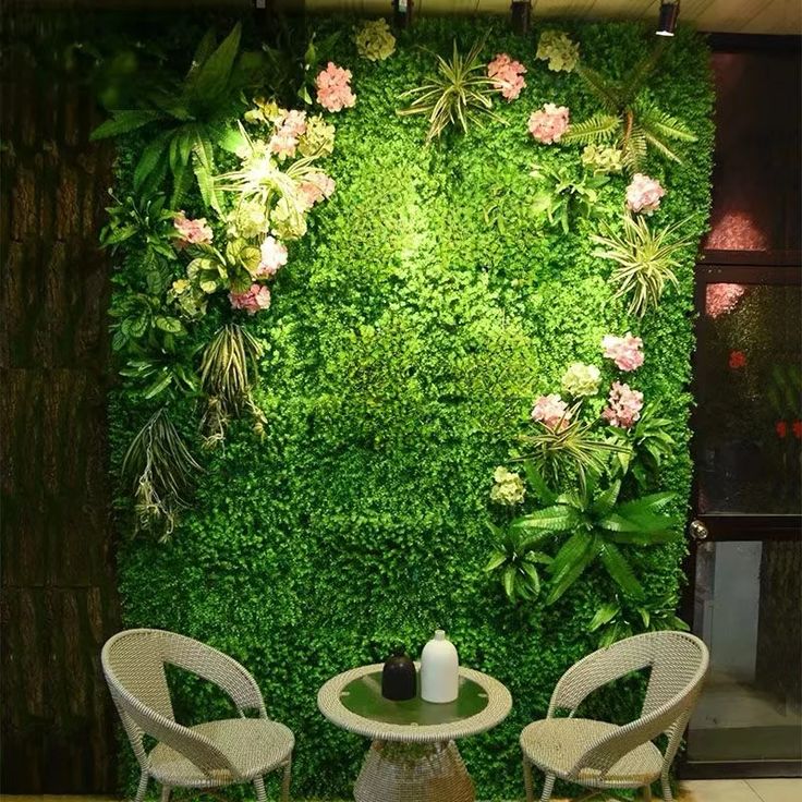 two white chairs sitting in front of a green wall with flowers and plants on it