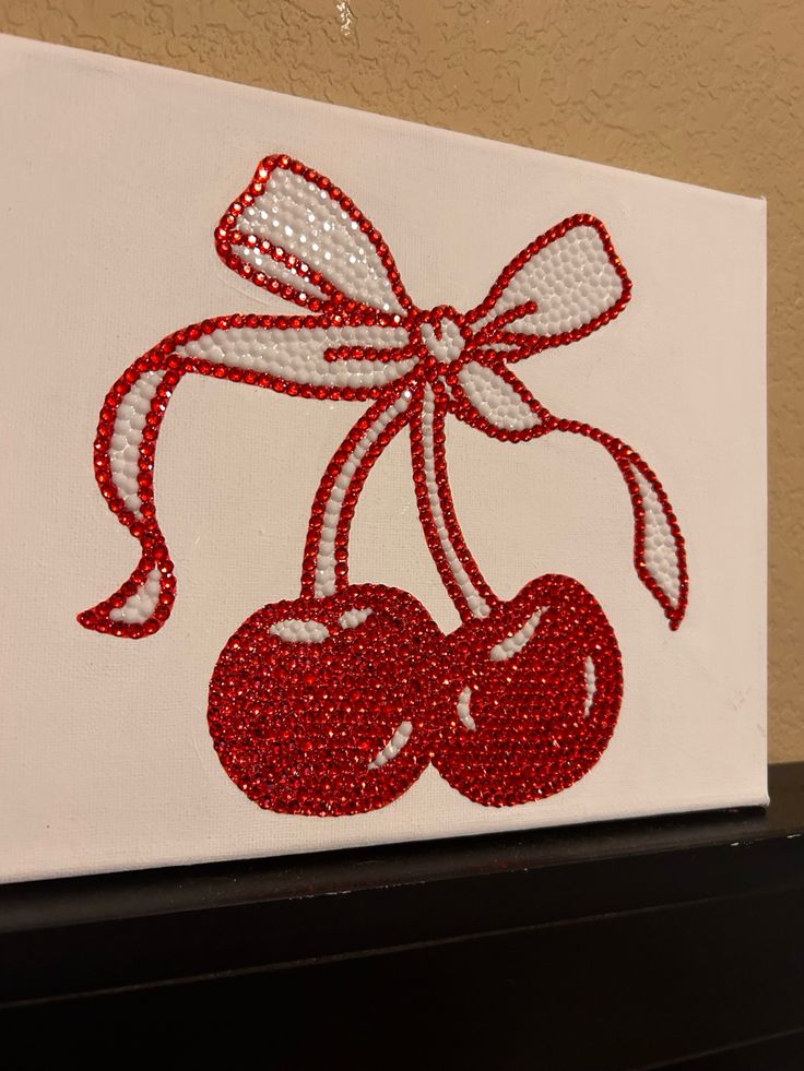 a white canvas with red and white sequins on it that has two cherries attached to the side