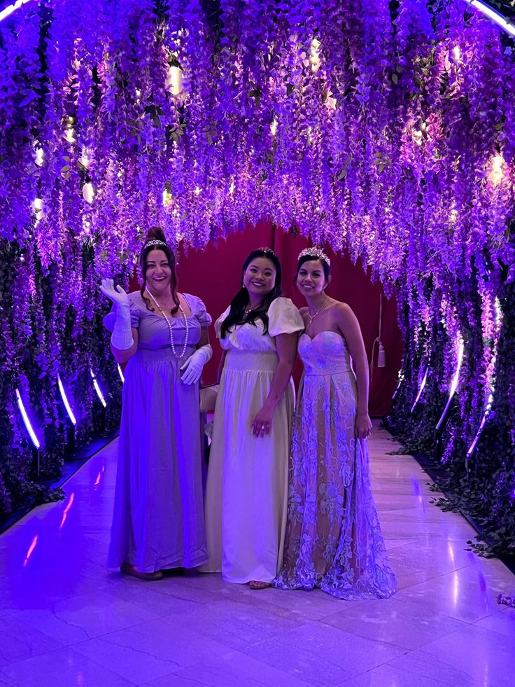 three women standing in front of a purple tunnel with wistery vines hanging from the ceiling