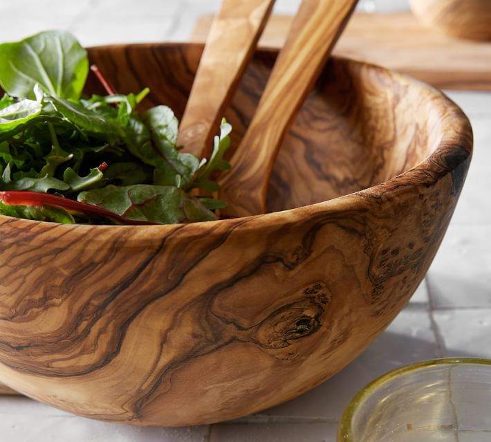 a wooden bowl filled with greens next to a bottle of olive oil and spoons