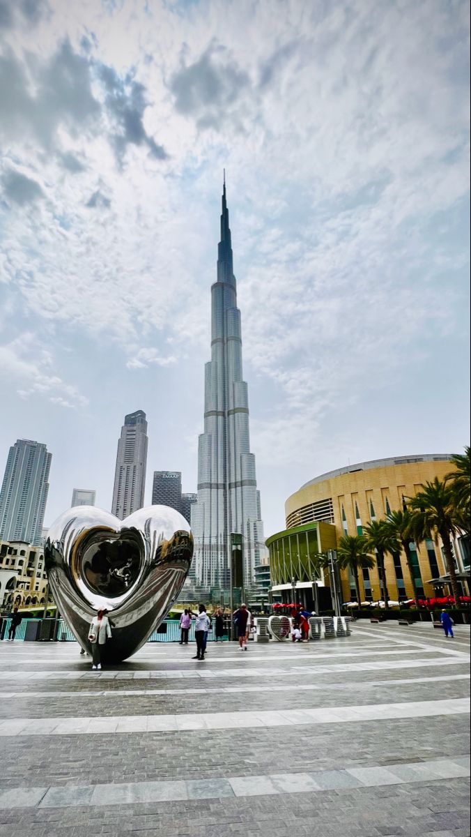 a large metal sculpture in the middle of a square with people walking around it and skyscrapers in the background