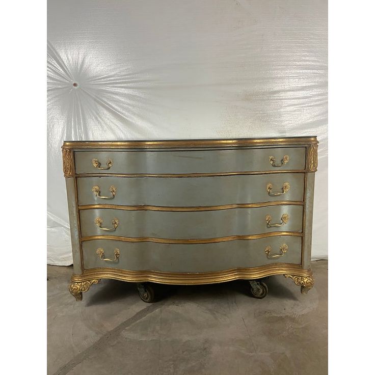 an antique chest of drawers with gold trimmings on the top and bottom, against a white wall