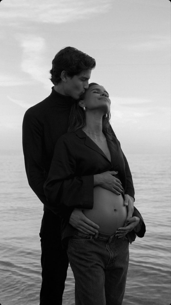 a man and woman standing next to each other near the ocean with their hands on their stomach