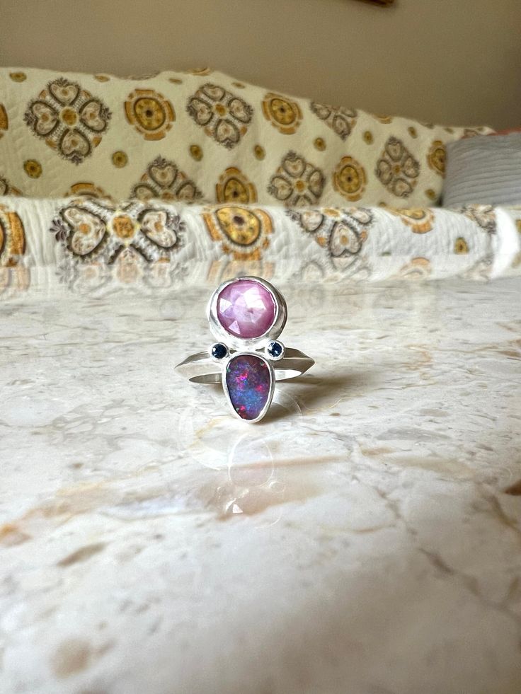 In bloom ring is set with a rose cut ruby, opal and two blue sapphires made in sterling silver. Opal has a pink and blue flash. Ruby has a wonderful opaque glisten and the cut gives it a little sparkle, nestled with the blue sapphires and opal it really sets it off.  US size 6 - can be sized up a bit Unique Pink Multi-stone Rings, Pink Sterling Silver Opal Ring For Anniversary, Pink Opal Ring In Sterling Silver For Anniversary, Pink Moonstone Sterling Silver Ring For Anniversary, Pink Sterling Silver Moonstone Promise Ring, Handmade Pink Ruby Ring In Sterling Silver, Pink Opal Sterling Silver Ring Gift, Pink Opal Ring In Sterling Silver For Gift, Unique Handmade Pink Ruby Ring