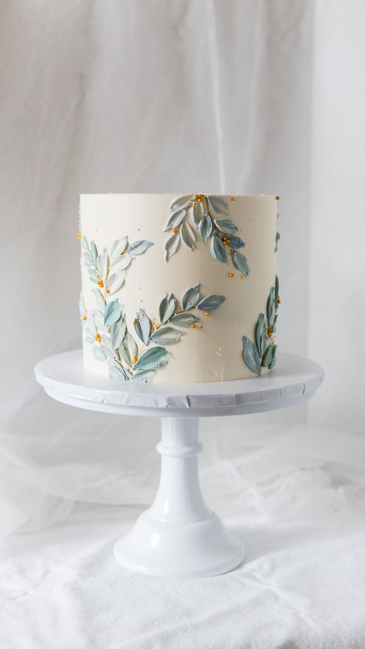 a white cake with blue leaves on it sitting on top of a table next to a white cloth