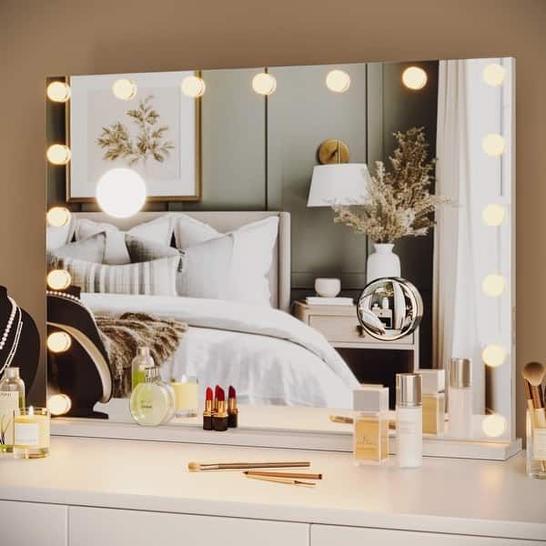 a vanity with lights and makeup on it in front of a mirror that is reflecting the bed