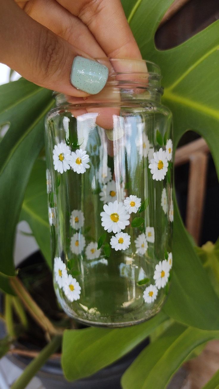 a person is holding a jar with flowers painted on it and there are green leaves in the background