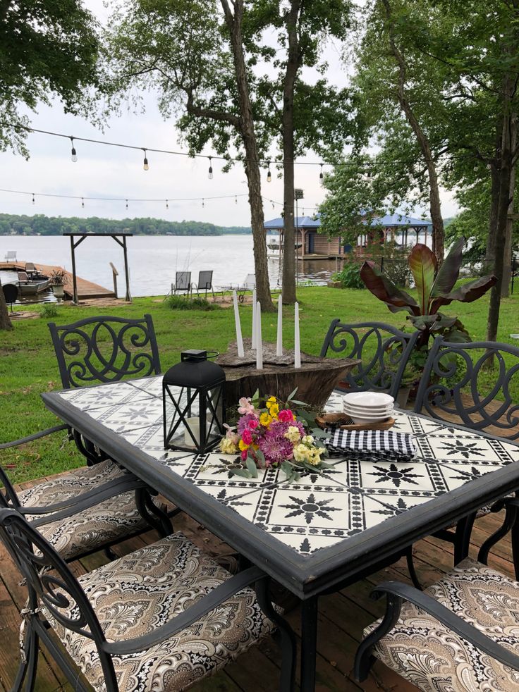 an outdoor table and chairs with flowers on it in front of a lake, surrounded by trees