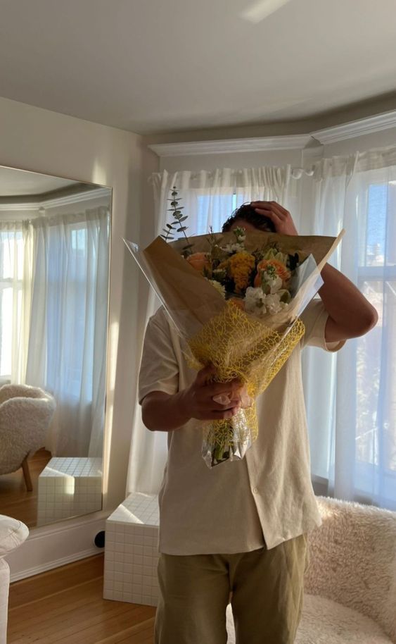 a man holding flowers in front of his face while standing in a living room next to a window