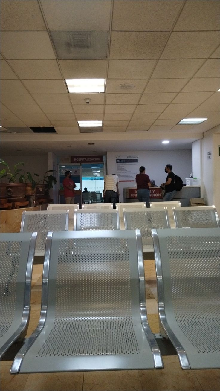 three empty metal seats in an airport waiting area with people standing around the seating areas